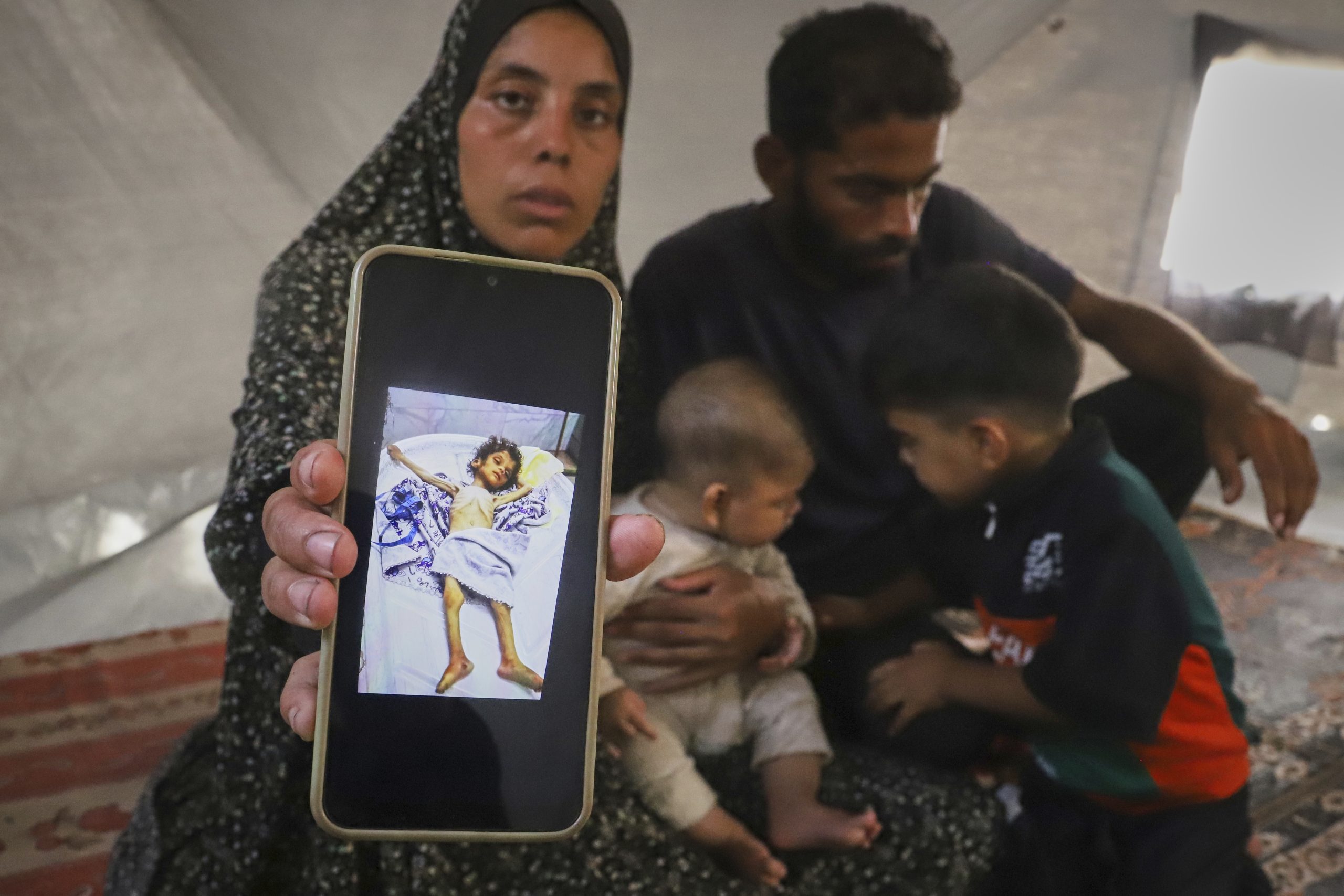 Sitting next to her husband, Amin, and two of their children, Fatma Mashi shows a photo on her cellphone of the body of her 2½-year-old daughter, Ro'a, who doctors say had no preexisting conditions and died of severe malnourishment, at their tent in Muwasi, southern Gaza Strip, Friday, Aug. 8, 2025. (AP Photo/Mariam Dagga)